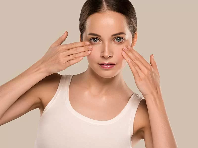 A imagem mostra uma mulher de pele clara e cabelos castanhos, presos em um coque. Ela veste uma regata bege e olha para a câmera. Suas mãos estão posicionadas abaixo de cada olho, deixando-os em evidência. O fundo da imagem é bege.