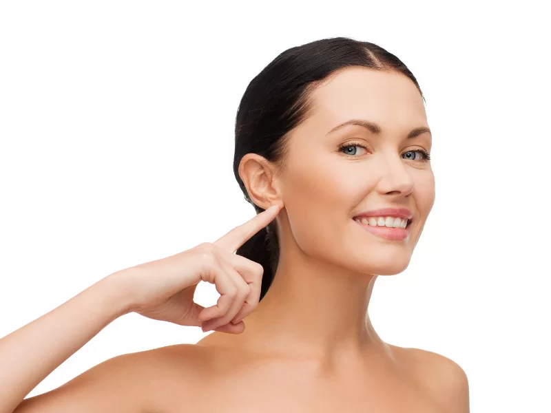 Imagem em ângulo oblíquo, que mostra uma mulher de cabelos pretos, em um quadro que pega da região do peito para cima, sorrindo ao encarar a câmera, e com o dedo indicador da mão direita apontando para o lóbulo da orelha direita.